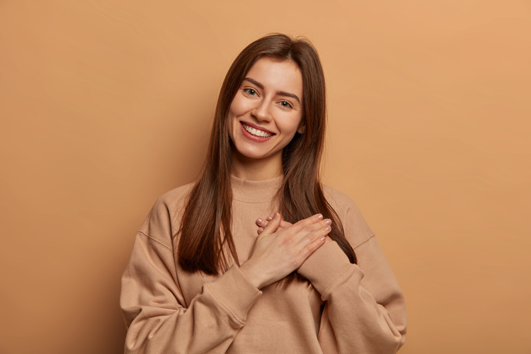 Touched pretty woman makes gratitude gesture, presses palms to heart, melts of adorable lovely gift received from friend, feels sympathy and passion, appreciates gift, stands against brown background